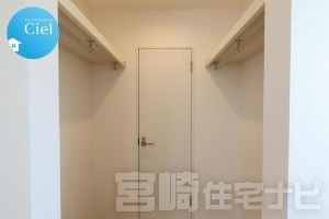 【ウォークインクローゼット】洋室1に併設された大容量のウォークインクローゼット。両サイドにハンガーパイプと枕棚を備え、ご夫婦で左右を使い分けたり、季節ごとに衣類を整理したりと機能的に使えます。最大の特徴は、奥の扉が室内干しに便利な「サンルーム」へと直結しているウォークスルー設計であること。サンルームで乾いた洗濯物を、たたまずにそのままハンガーにかけて収納できる、究極の「家事ラク動線」を実現しました。足元の空いたスペースには、衣装ケースや旅行用のトランクなどのかさばる荷物もすっきり収まり、お部屋を広々と保つことができます。